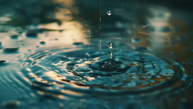 A water drop sending ripples across blue-green water, with warm golden reflections.