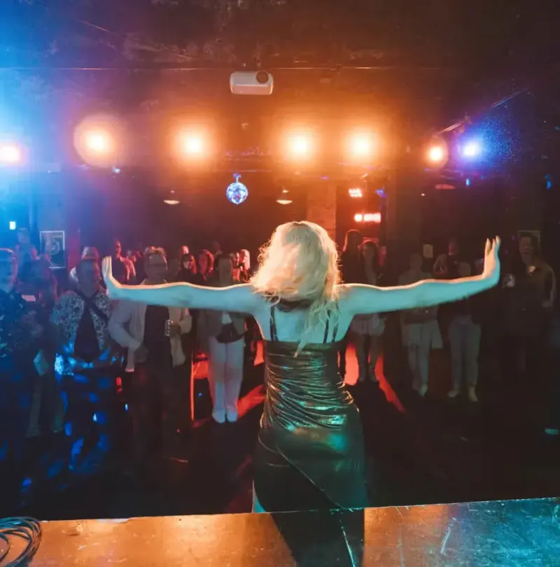 Performer in a sparkly dress with arms outstretched facing a lit crowd on a club stage at the retreat
