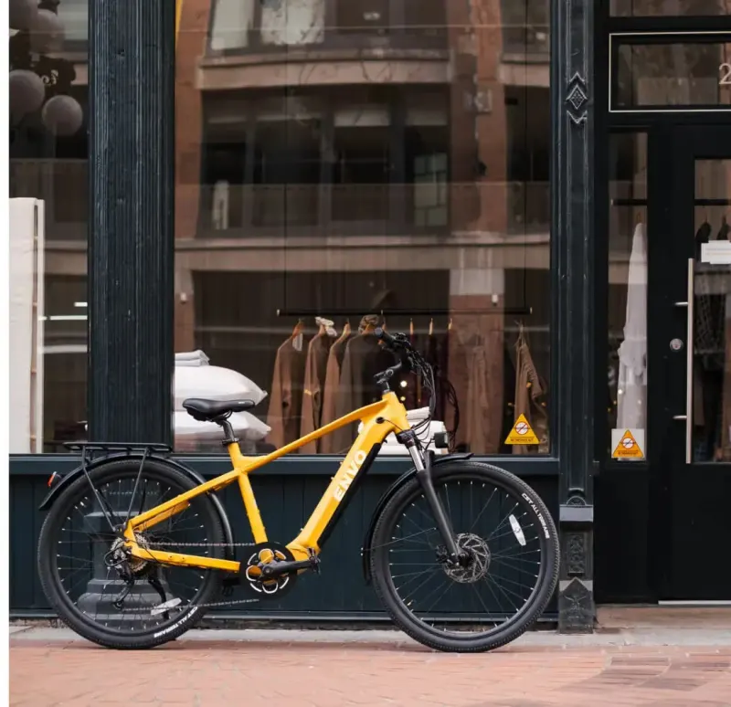 Yellow ENVO e-bike leaning against a dark heritage storefront with a clothing display on a brick sidewalk