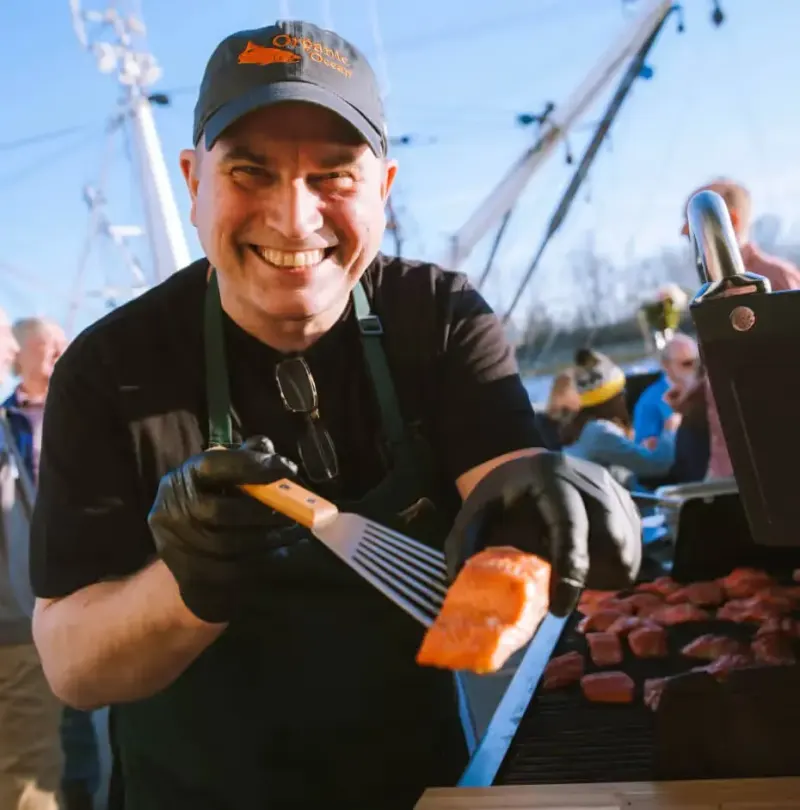 Smiling grill cook in an Organic Ocean cap holding a salmon fillet on a spatula at a waterfront event