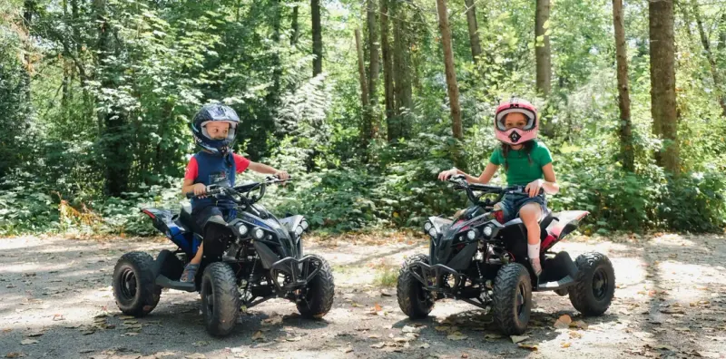 Two children in helmets riding Rosso youth ATVs side by side on a wooded gravel path