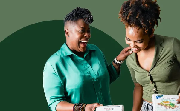 Two women laughing together over a package on a green ground