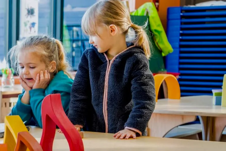 Two young girls at a classroom table with red and yellow wooden blocks, one resting her chin in her hands