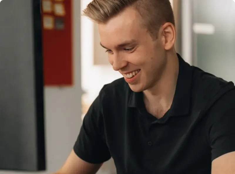 A young man in a black polo smiling at his desk