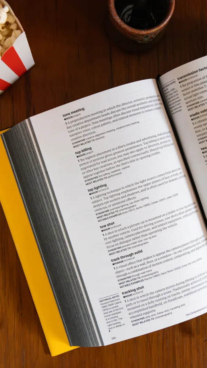 An open book of film terms on a wooden table beside a bucket of popcorn and a cup of coffee