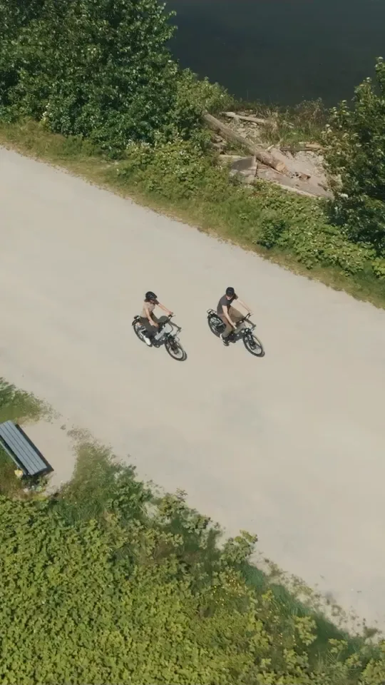 Two riders on e-bikes along a gravel path beside the water, filmed from above