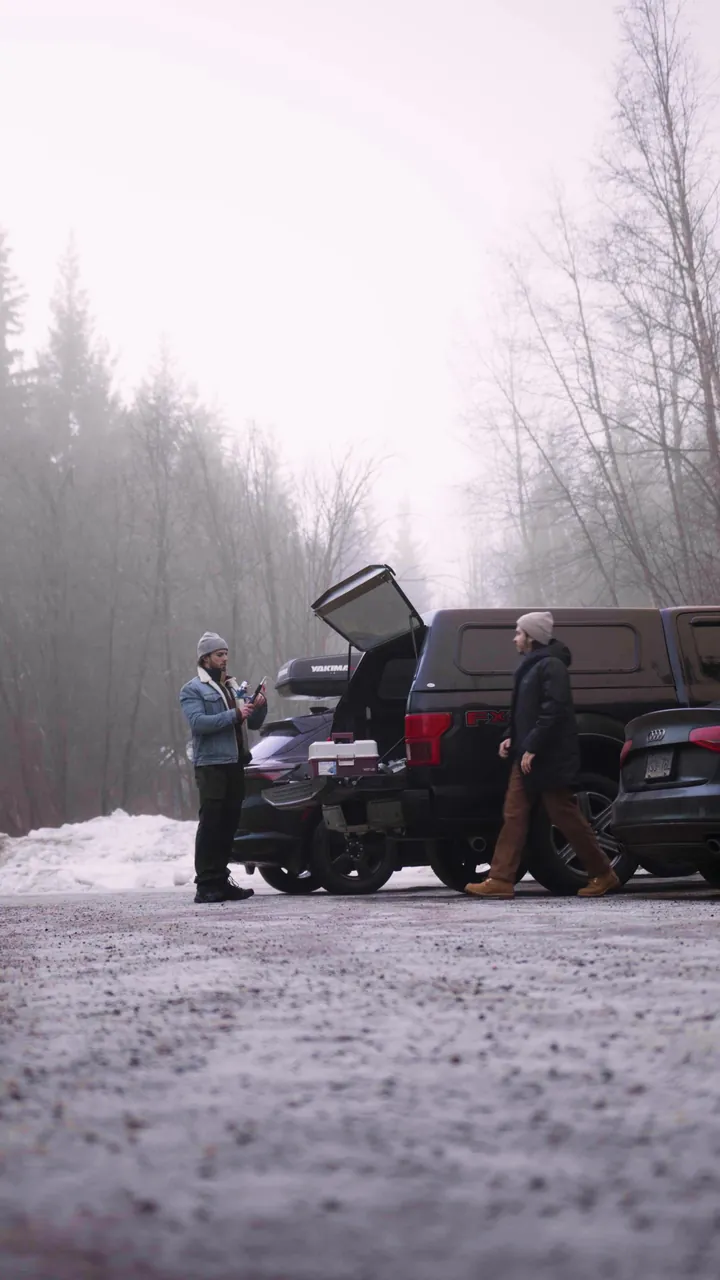 Two men loading gear into the back of a pickup on a snowy road, in fog among bare trees