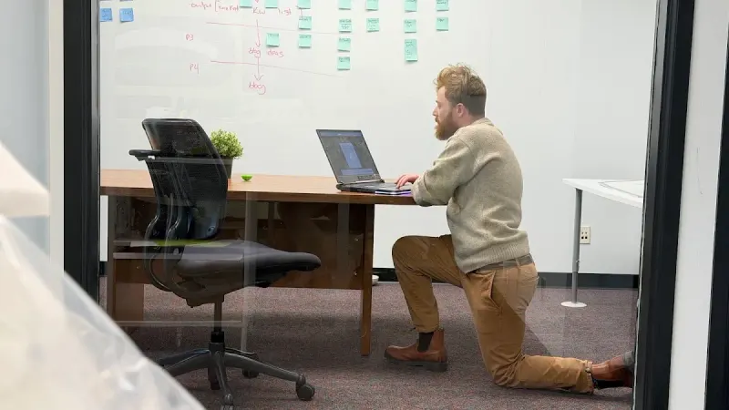 A Setsail team member kneeling at a desk to work on a laptop in a glass-walled office, a whiteboard of sticky notes behind him