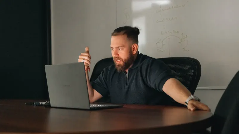 A bearded Setsail team member talking through an idea at his laptop, at a round table with a whiteboard behind him