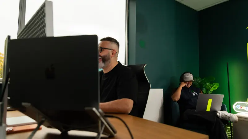 A Setsail team member smiling at his laptop at a desk, a colleague working on a laptop on the couch behind him in a green-walled room