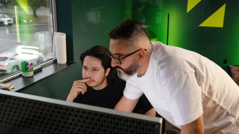 Two Setsail team members looking at a monitor together, one leaning over the other’s shoulder, in front of a green wall