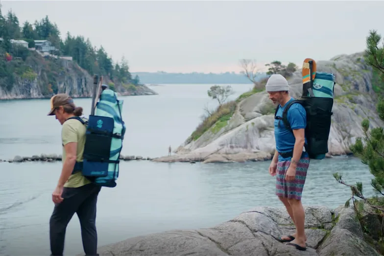 Two people carrying Squid Paddle Co. board packs along a rocky shoreline