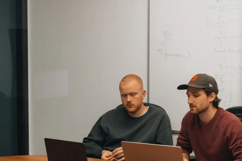 Two Setsail strategists at their laptops in front of a whiteboard, one in a cap