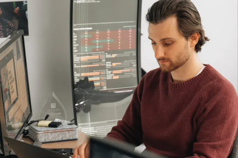 A strategist in a red sweater working at his desk beside a monitor of market data