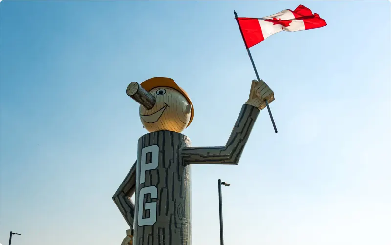Mr. PG, Prince George's log mascot statue with a hard hat, waving a Canadian flag against a blue sky
