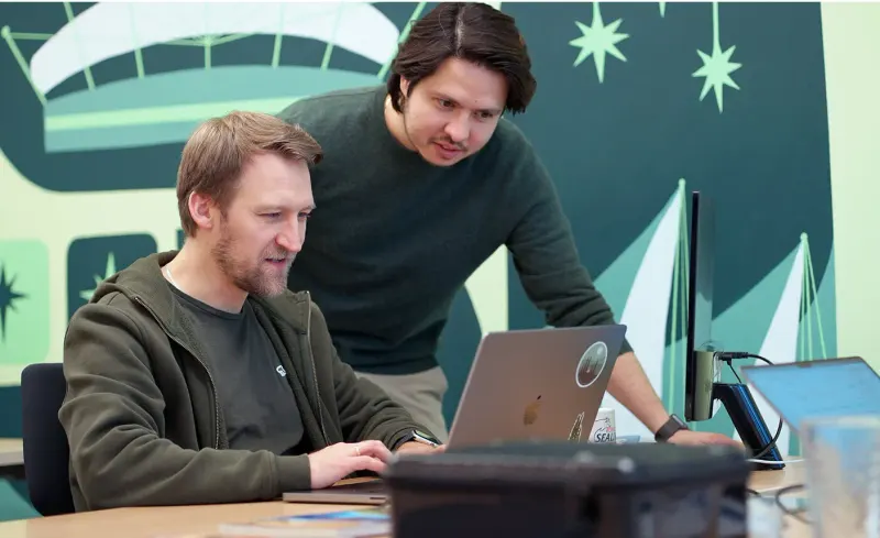 Two Setsail team members reviewing work on a laptop in an office with a teal illustrated mural behind them