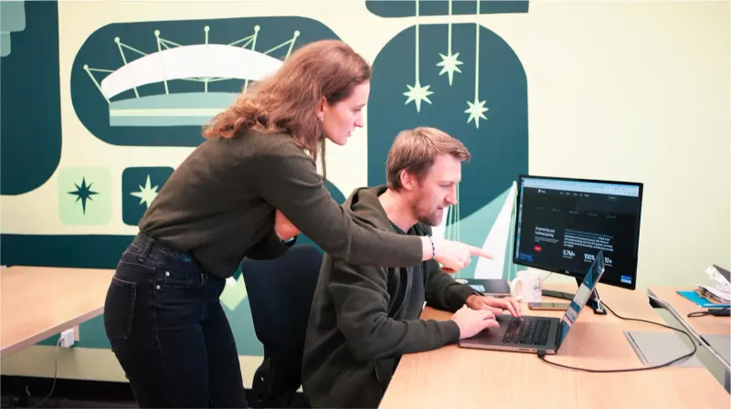 Two Setsail team members at a desk reviewing a website on a laptop and monitor in front of a colourful wall mural