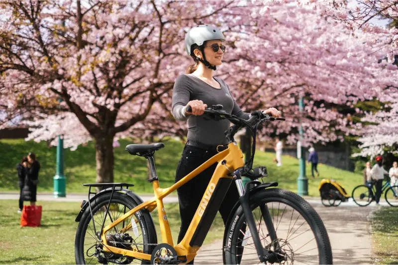 Woman in a helmet and sunglasses standing with a yellow ENVO e-bike beneath pink cherry blossom trees in a park