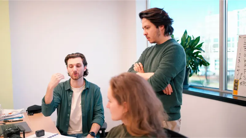 Three team members in a bright office discussing a project, one gesturing while another stands with arms crossed