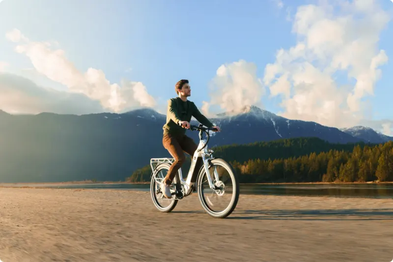 A rider on a white DŌST e-bike on a beach, mountains behind