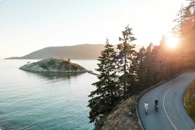 Two cyclists on a winding coastal road above the sea at sunset