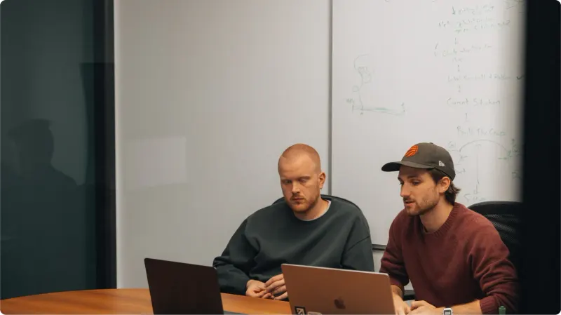 Two men working at laptops at a meeting table in front of a whiteboard