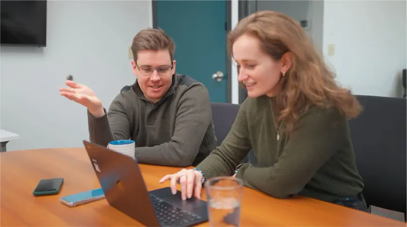 A man and woman seated at a conference table discussing work on a laptop