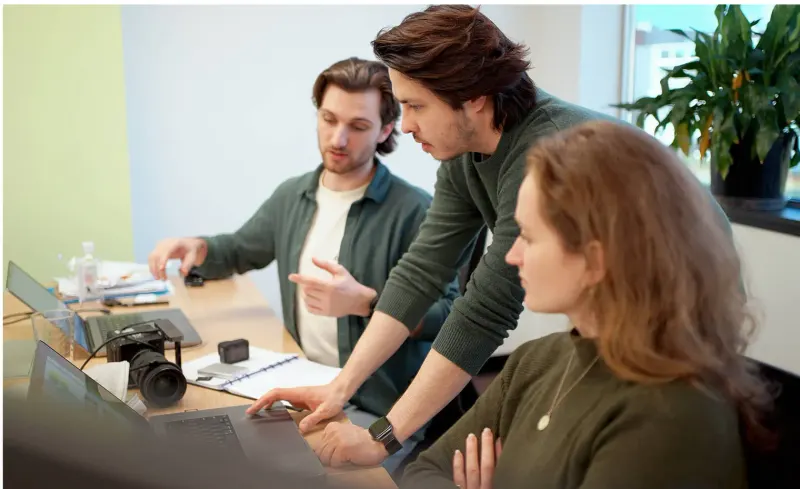 Three team members reviewing work on laptops at a shared desk, with a camera and notebook on the table